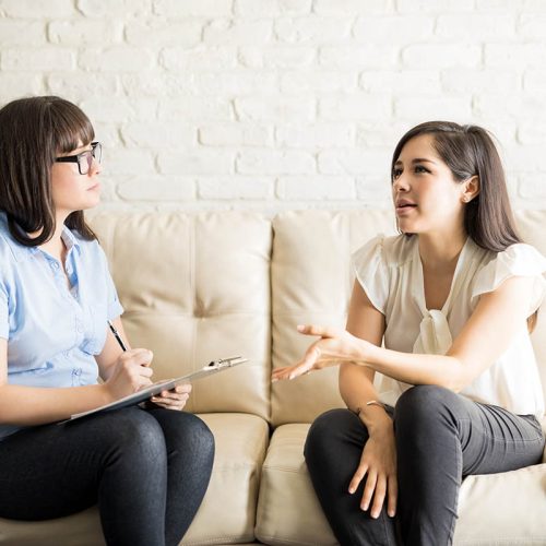 Pretty young woman talking to female therapist on sofa, sharing her issues during psychotherapy.